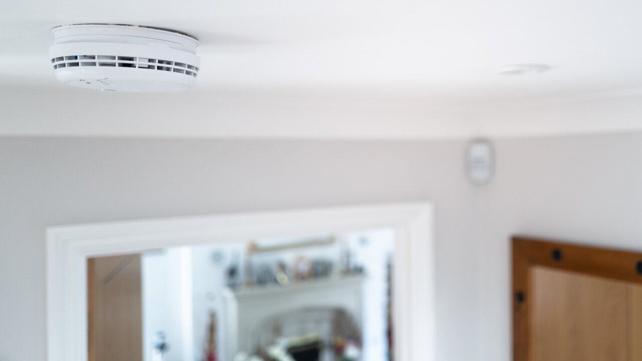 White smoke detector mounted on a home ceiling near a hallway and fireplace.